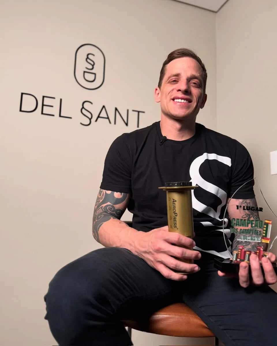 Mayonn sorrindo, segurando uma AeroPress e o troféu de campeão dos campeões